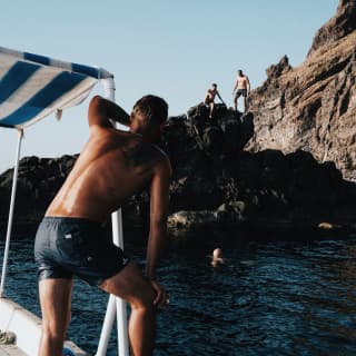 A tanned man, wet from a dip in the sea, leans on his boat's canopy bar as he chats to friends on the cliff and in the sea