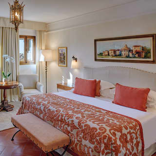 A painting of a Tuscan village hangs above the bed head. Cream upholstered chairs sit on a cream rug on the terracotta floor