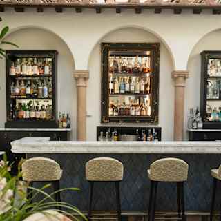 Four stools line a white marble bar with teal front facing three gleaming bottle shelves spaced between decorative wall arches.
