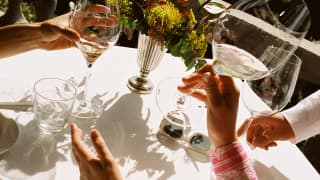 Close-up of an outside table at La Loggia where a guest wearing long pink sleeves raises a glass of white with two friends.