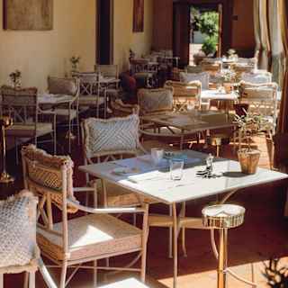 View through a sun-room style dining area with rows of tables, half in sunshine, and cushioned chairs, set on terracotta tiles.