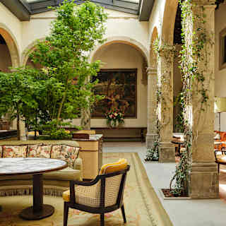 A beige sofa with floral cushions, a cane chair and marble table sit on a vintage rug in a cool bright cloistered courtyard.