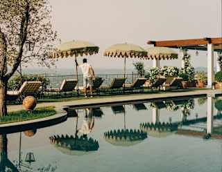 A man adjusts one of three sage-green parasols shading a row of chairs with hilltop views, mirrored in the calm pool waters.