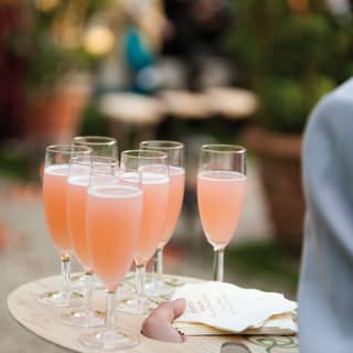 A waiter holds seven Bellinis in champagne flutes served on a tray made from an artist's wooden paint palette