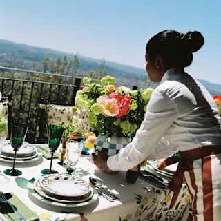 A female staff member places a vase of green and pink ranunculi on a sunny, view-filled terrace lunch table, seen from behind.
