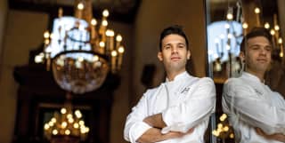 Alessandro Cozzolino, leans on the edge of an open glass door. His folded arms and chef whites reflected in the glass