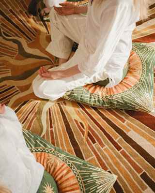 A woman in white, seen from the shoulders down, sits in meditation pose on a cushion and throw in browns and greens.