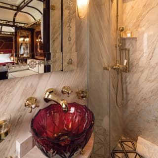 Grey-veined white marble covers every surface of this extravagant bathroom. A gold tap curves over a red glass bowl sink