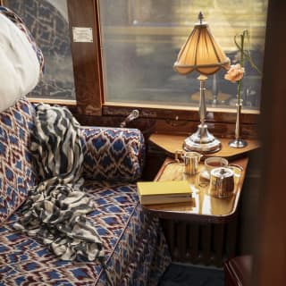 A lamp, a pot of tea and a book rest on a table in a cabin interior, with upholstered banquette in rich blues and maroons.