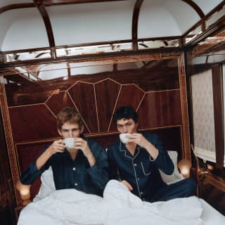 Two men in navy pyjamas sip coffee beneath the white duvet of their bed in the Prague Suite, photographed by Coco Capitán.
