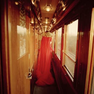 Lady in a vibrant floaty red dress strolling away from the camera down a wood-panelled train carriage