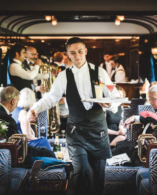 Barman holding cocktails on a tray and stepping through a train bar car