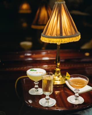 Below the golden glow of a silk shaded table lamp, two cocktails and a glass of champagne sit on a polished table