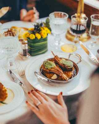 Restaurant detail as a woman rests her hand on a dining table next to a silver bowl with two herb-crusted lamb chops.