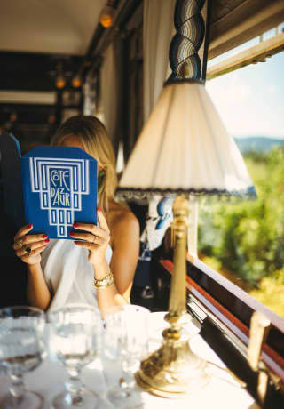 Seen across a table with a lamp and three glasses, a woman holds a blue Cote d'Azur Dining Car menu in front of her face.