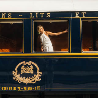 A blond woman in a white halter-neck dress stretches an arm across the train window as she peers out, seen from the platform.