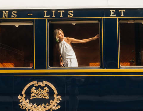 A blond woman in a white halter-neck dress stretches an arm across the train window as she peers out, seen from the platform.