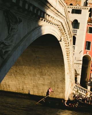 Under the Rialto Bridge, a gondolier in a red and white striped top uses a rèmo to steer his boat, invisible in the shadows.