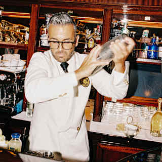 A barman in a white suite with black tie and spectacles shakes a cocktail behind a gleaming wooden bar, packed with bottles.